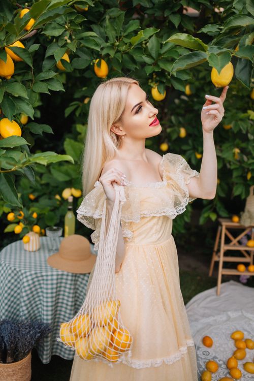 Free Young woman in a yellow dress picks lemons from a tree in a sunny orchard. Stock Photo