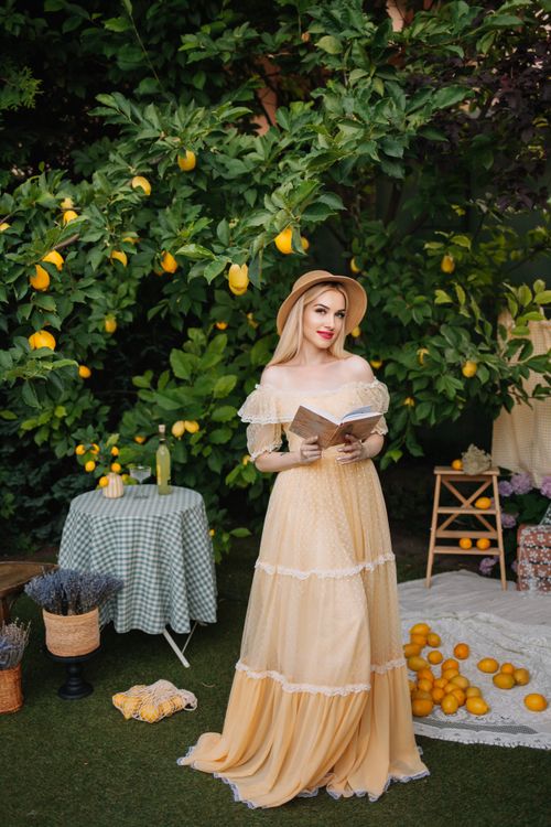 Free A woman in a flowing dress reading a book in a vibrant garden with lemon trees. Stock Photo