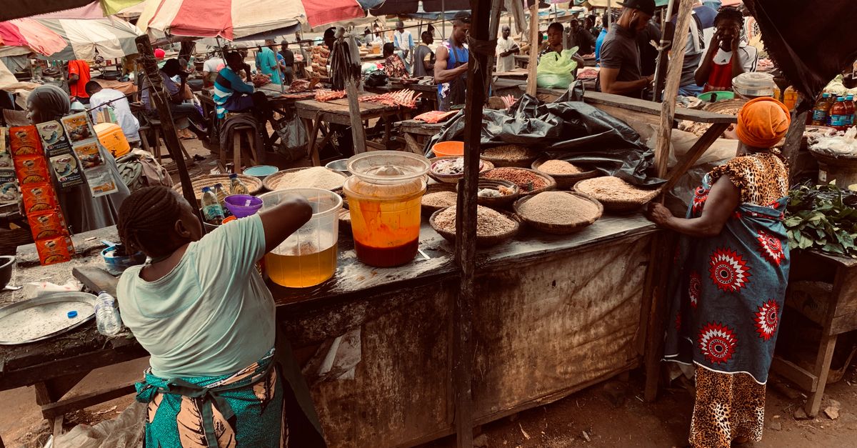 A bustling street market outside a church in Ho, Ghana, with women selling food from wooden stalls, steam rising from pots, and colorful cloths covering tables