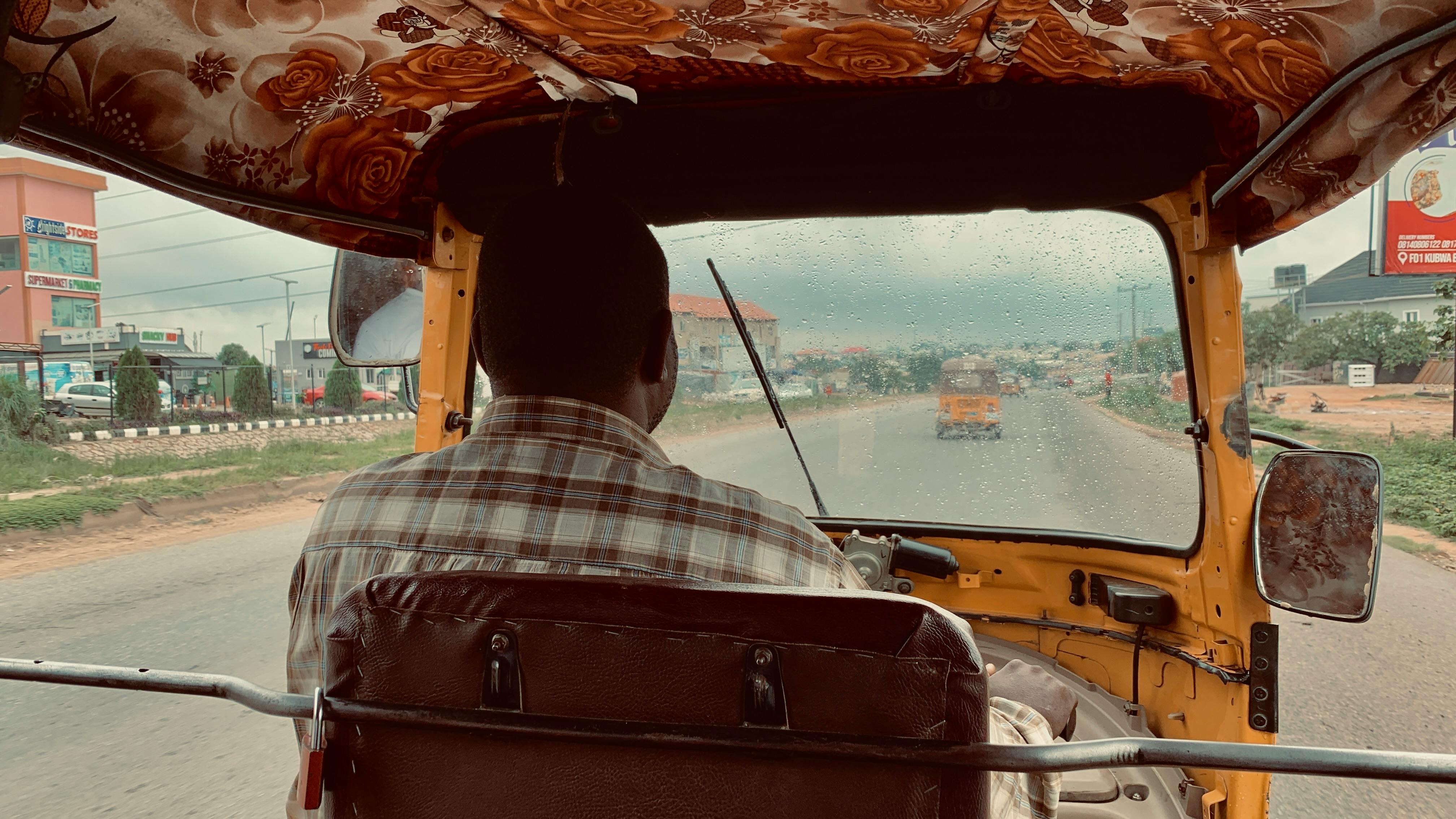 A man drives an auto rickshaw on a rainy street through an urban area.