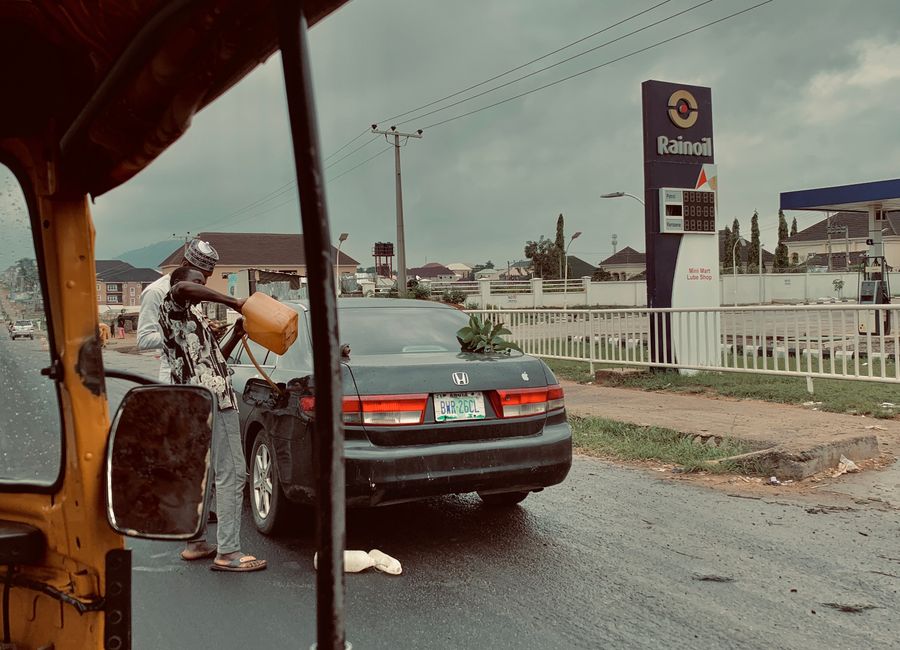 A man fills petrol from a container into a car by a Rainoil station, on a rainy street.
