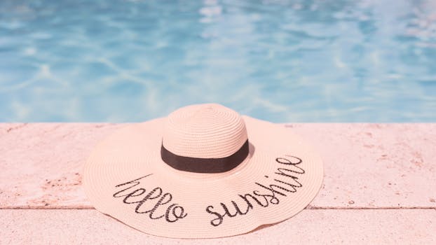 A straw hat with 'Hello Sunshine' embroidery by a sparkling swimming pool on a sunny day.