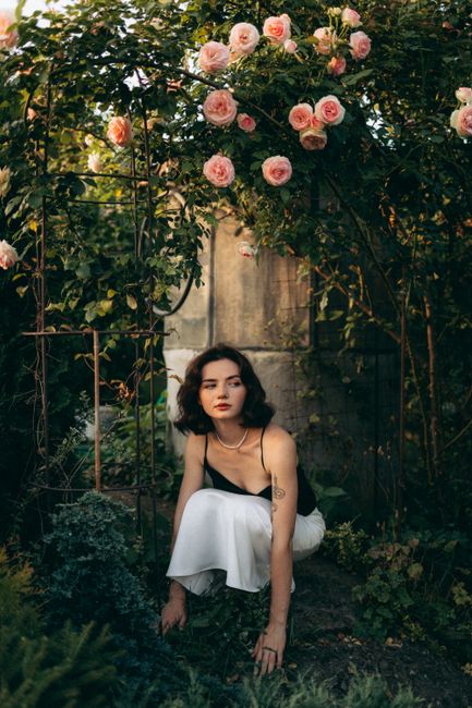 Elegant woman posing amid blooming roses in a lush garden setting.