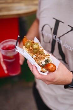 Person holding a gourmet hot dog with jalapeños and a refreshing drink outdoors.