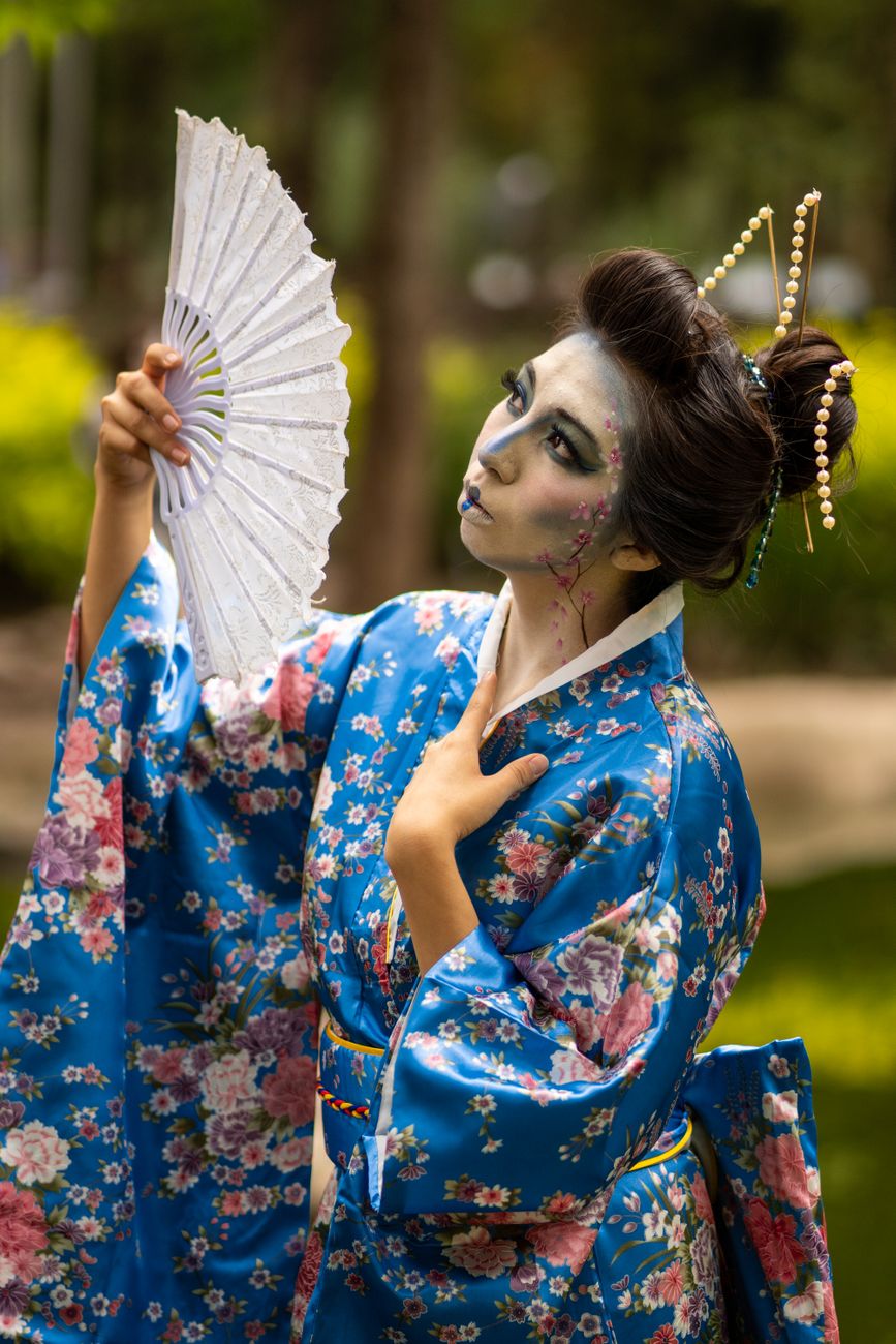 A vibrant portrait of a woman in a floral kimono and creative makeup holding a fan outdoors.