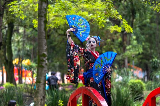 A woman in geisha attire with blue fans poses on a bridge in a lush park in Mexico City.