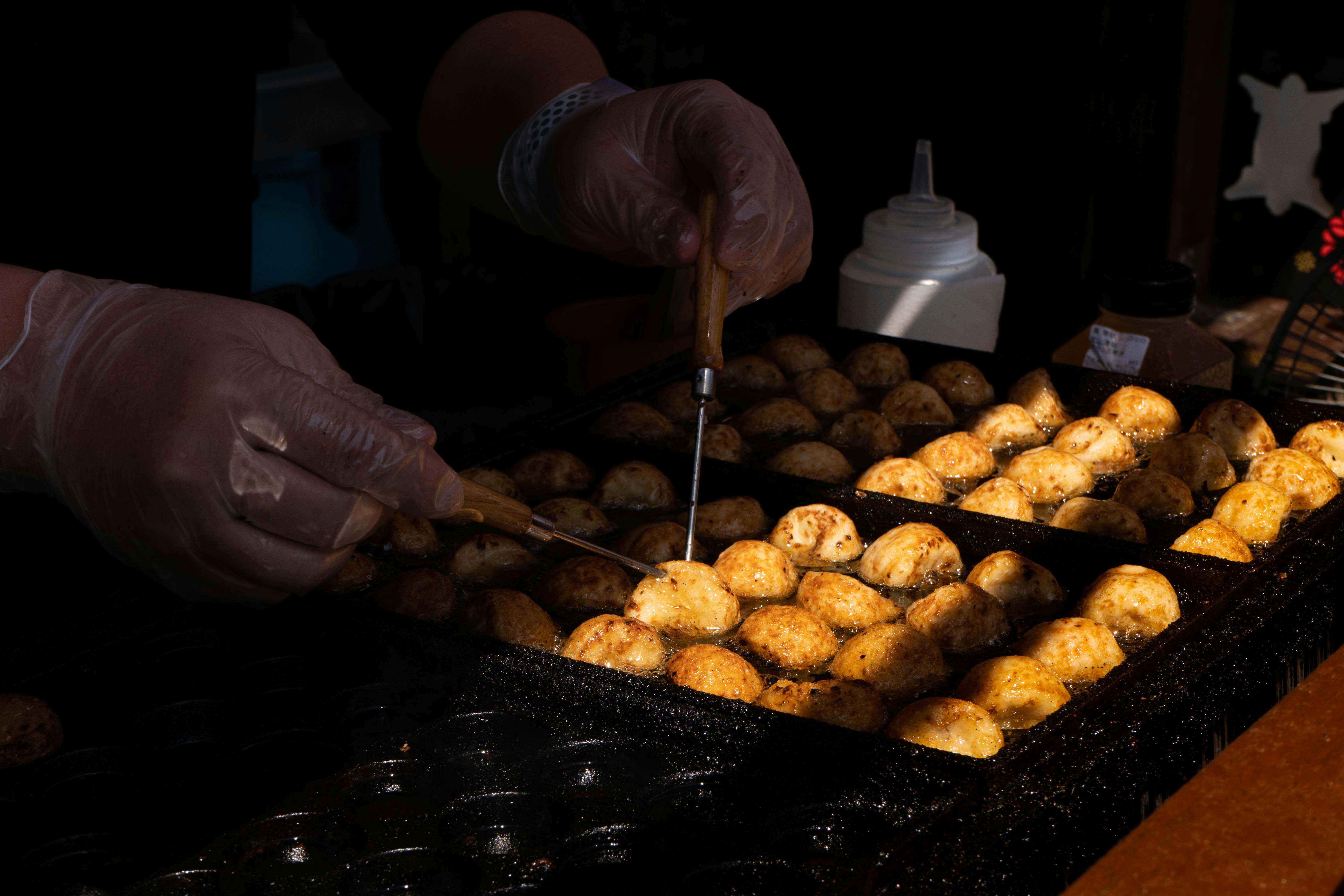 Hands turning takoyaki balls on a hot griddle at a Japanese street food stall.