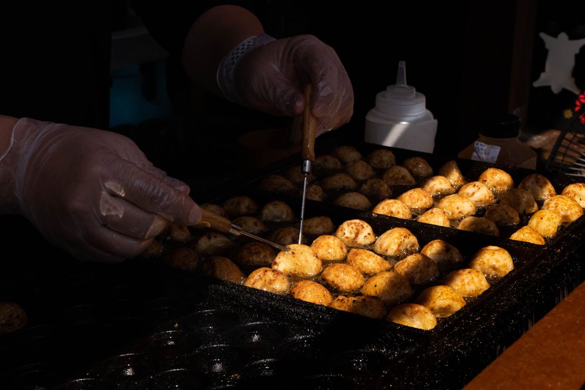 Gloved hands turning half-cooked takoyaki with two thin picks on a wide iron griddle, the balls glowing gold where the heat has caught them.