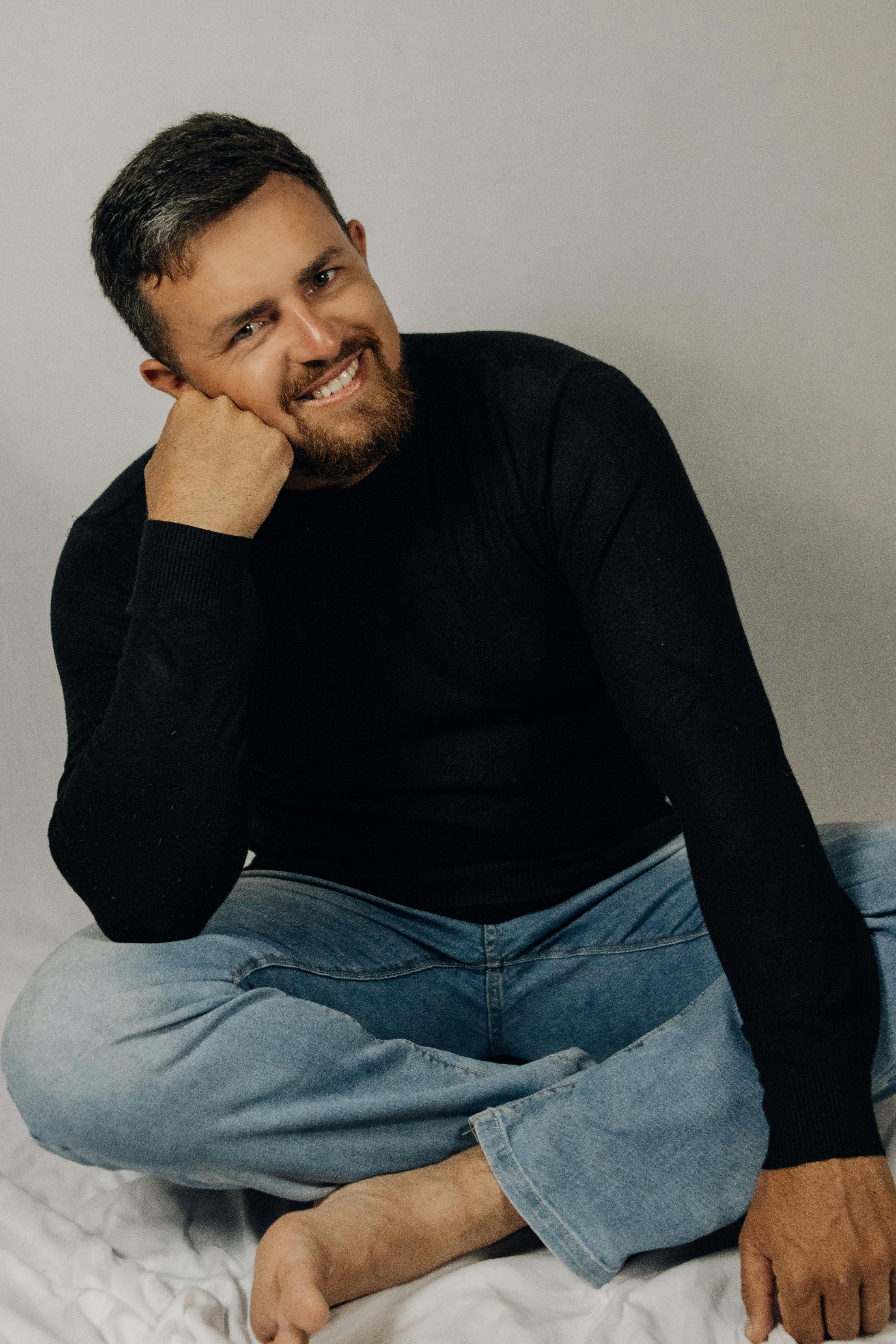 a man in a black sweater and jeans sitting on a bed