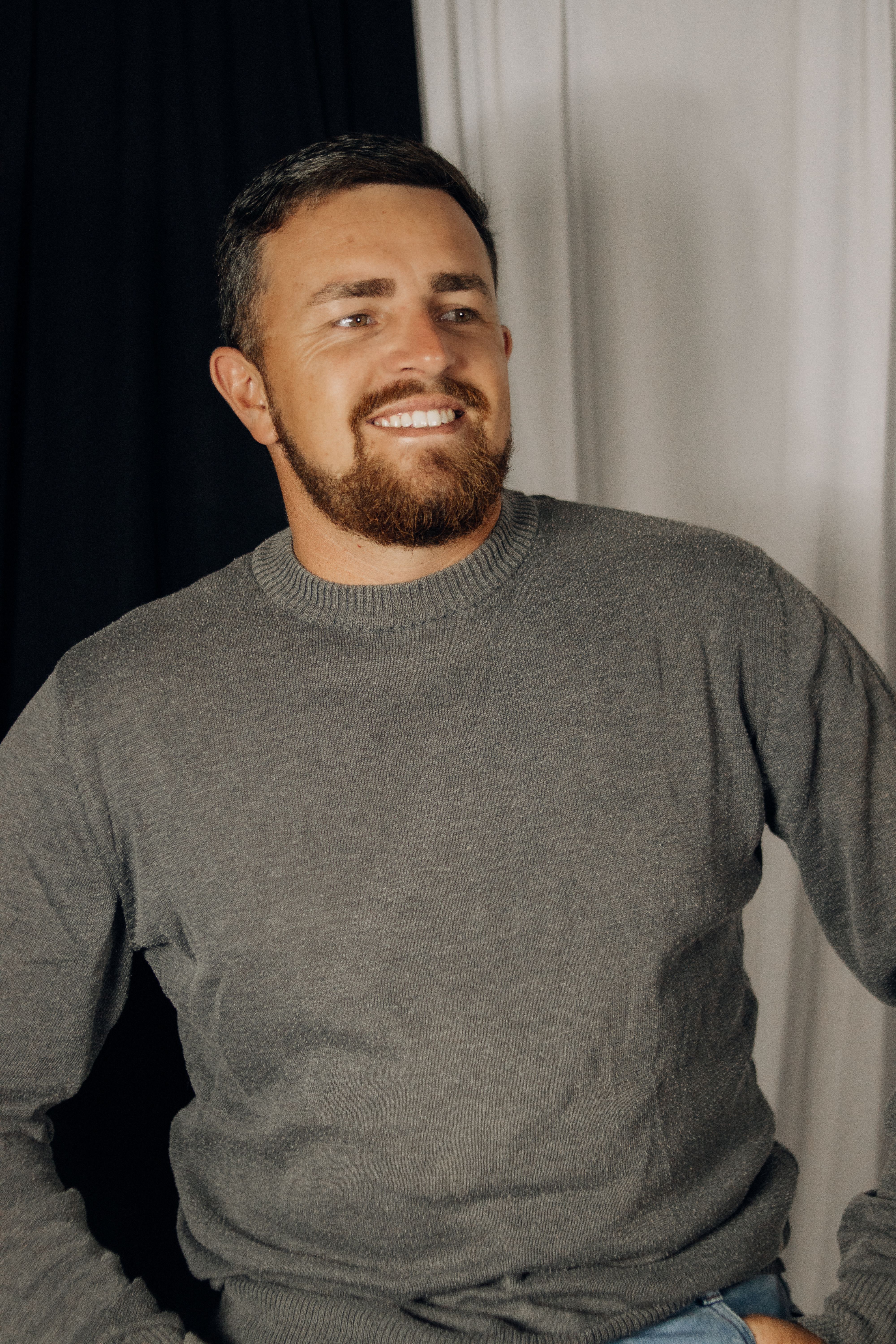 Portrait of a smiling man with a beard and casual attire, set in a studio.
