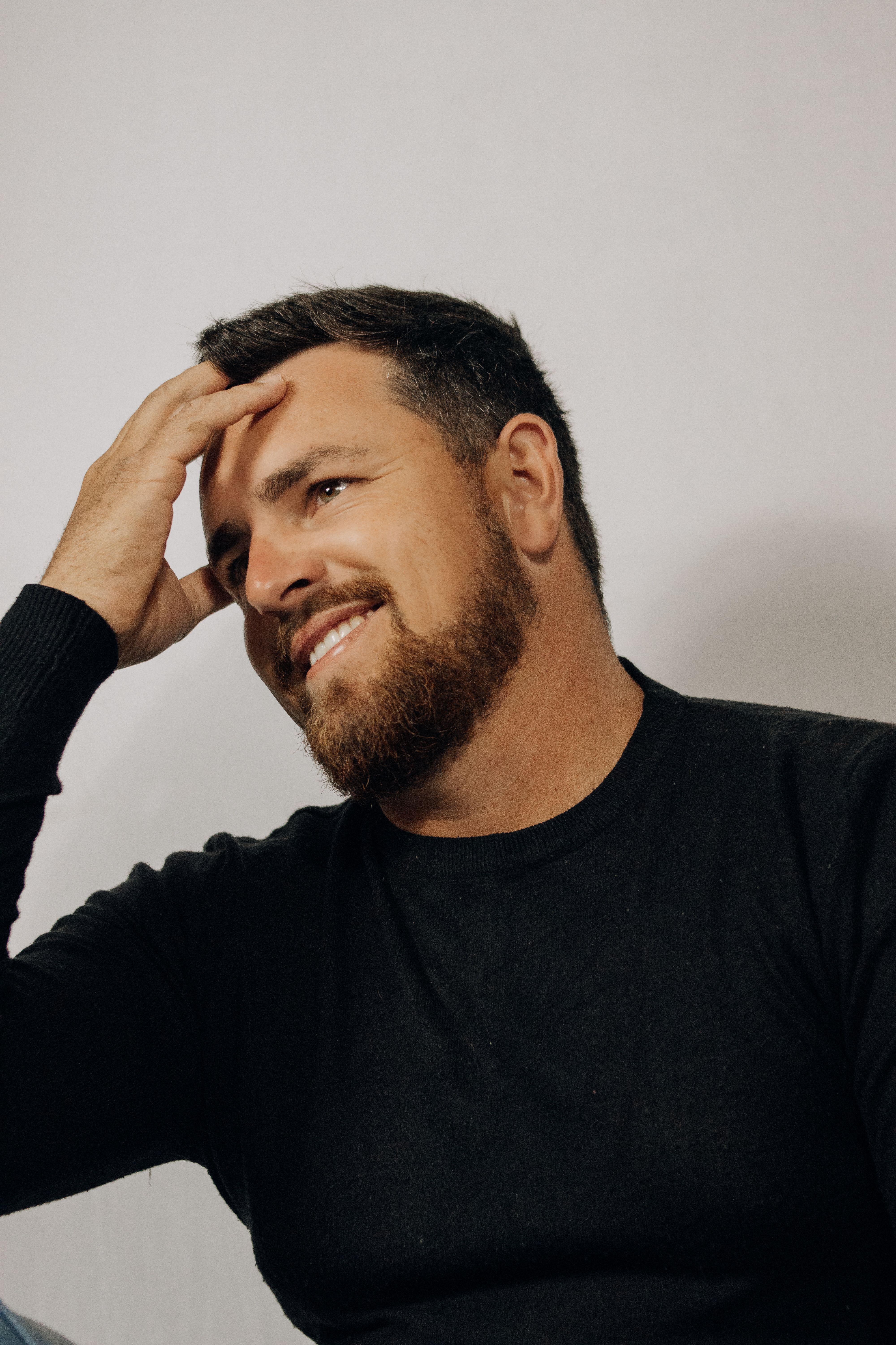 A thoughtful man with a beard and black sweater, posed indoors against a simple background.
