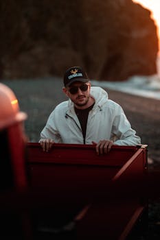A man wearing a white jacket and sunglasses stands by a beach at sunset, offering a moody vibe.