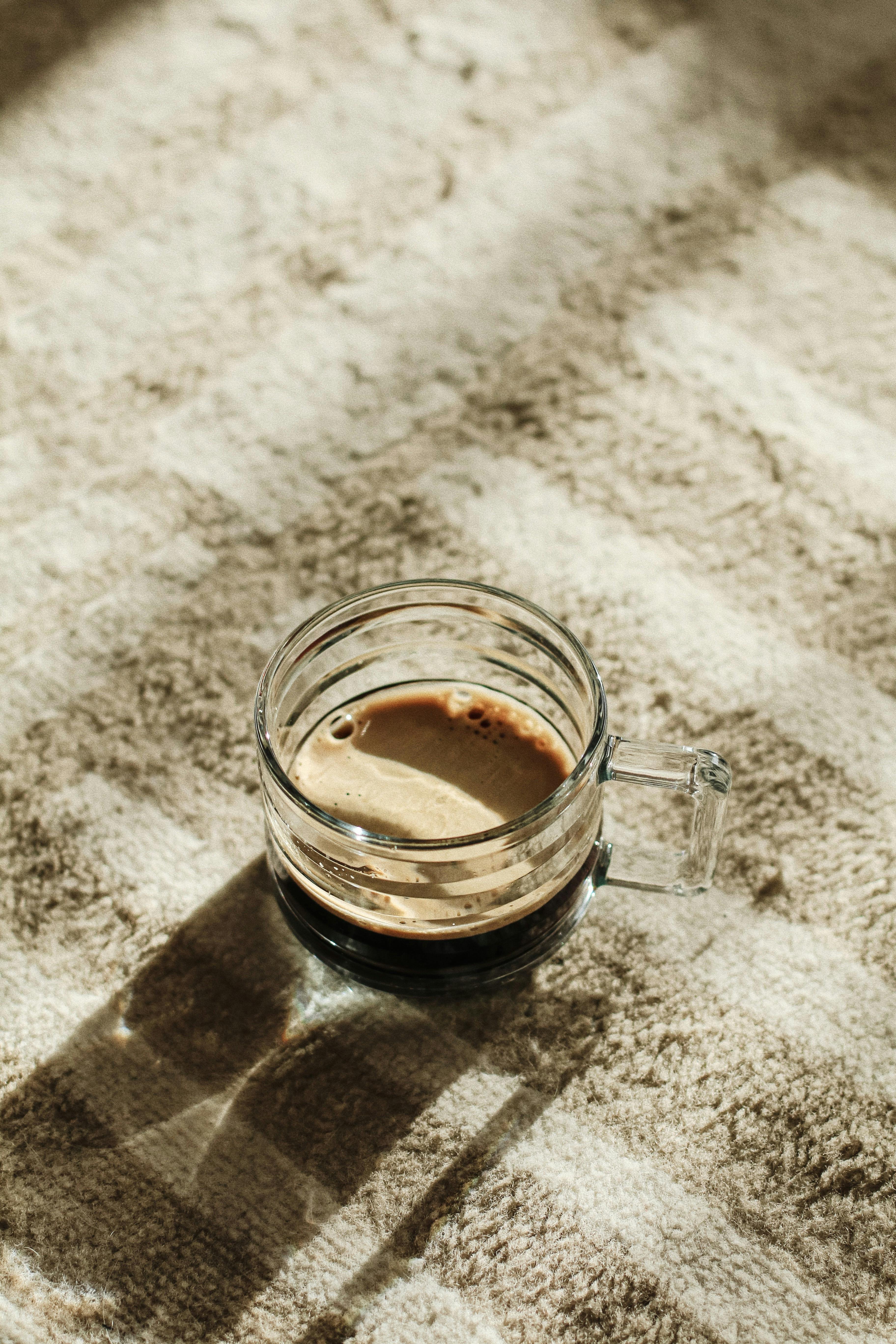 A clear glass cup of espresso casts a shadow on a textured beige surface, with warm lighting.