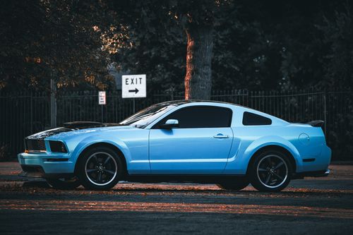 Free A classic blue Mustang coupe parked on a city street, highlighted by evening light and surrounded by trees. Stock Photo