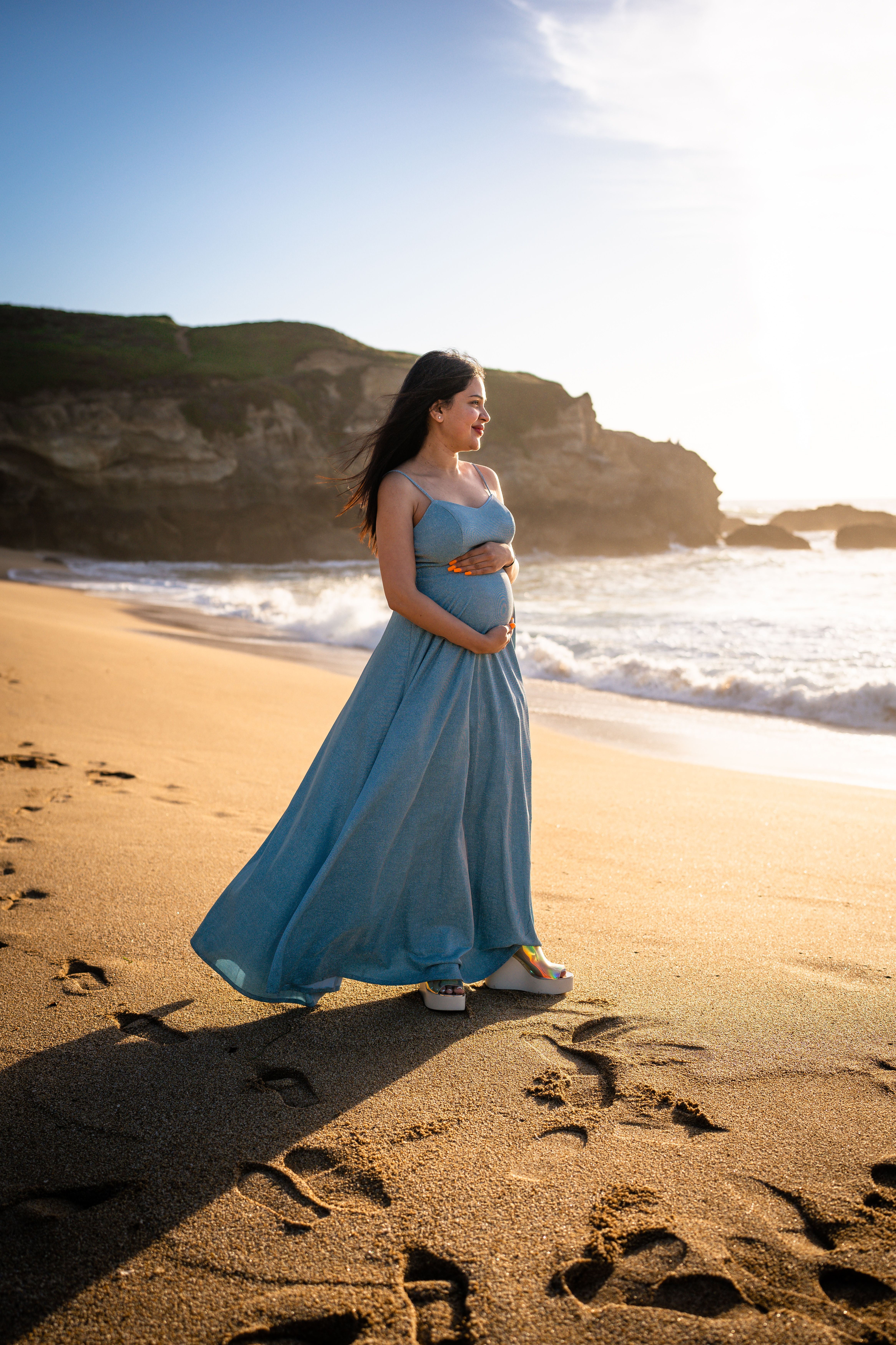 A pregnant woman in a blue dress stands on the beach · Free Stock Photo