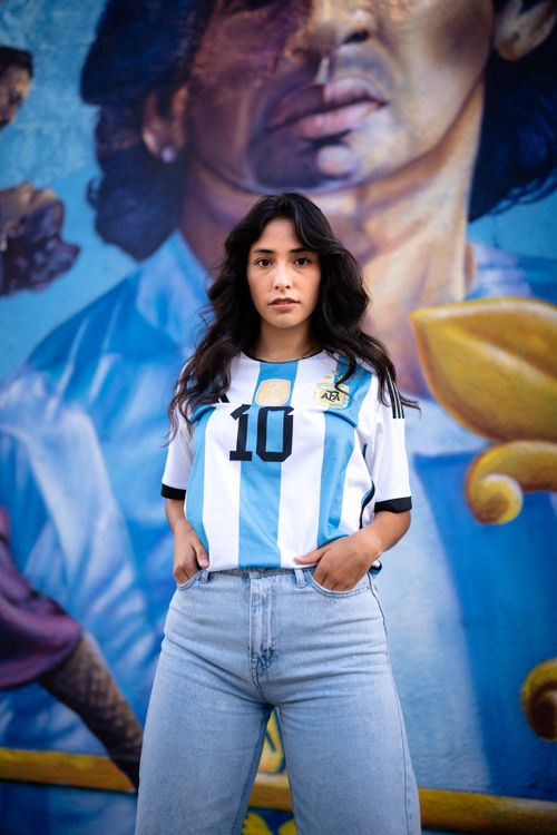 Free Young woman in Argentina jersey poses against vibrant mural backdrop capturing urban culture. Stock Photo