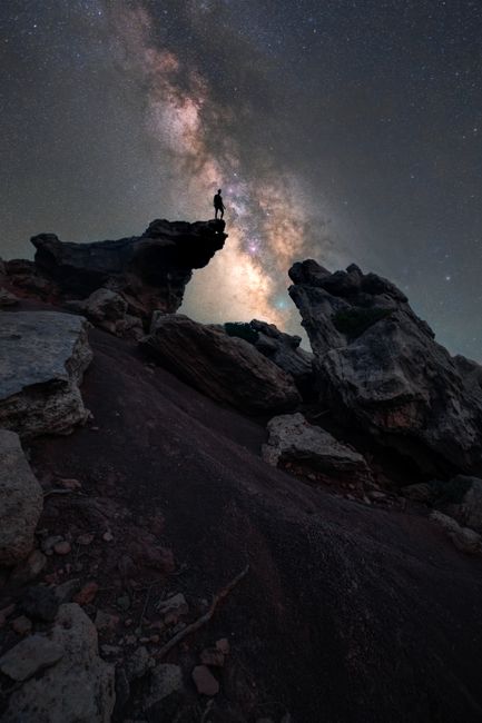 A lone figure stands on a cliff silhouetted by the Milky Way, capturing a breathtaking night sky.