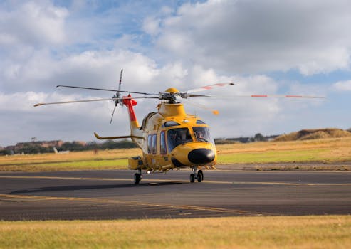 A yellow helicopter on an airfield ready for takeoff under a cloudy sky.