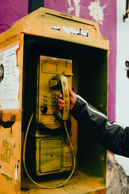 Close-up of a retro yellow phone booth with graffiti in an urban setting. Hand holding the receiver.