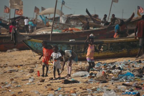 Free Children play amidst trash on a polluted beach with fishing boats in the background. Stock Photo