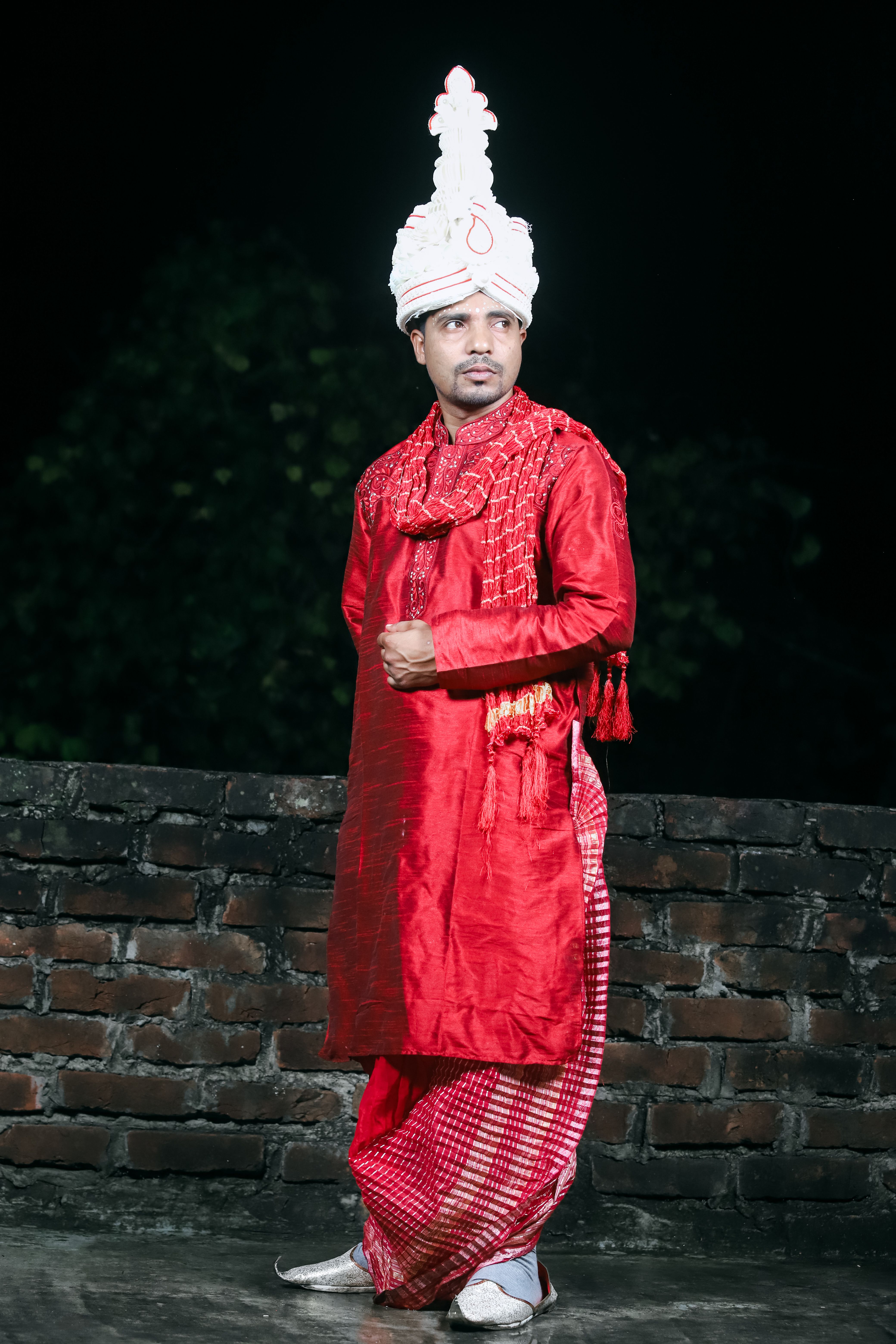 A man in traditional red attire with a white turban poses against a brick wall at night.