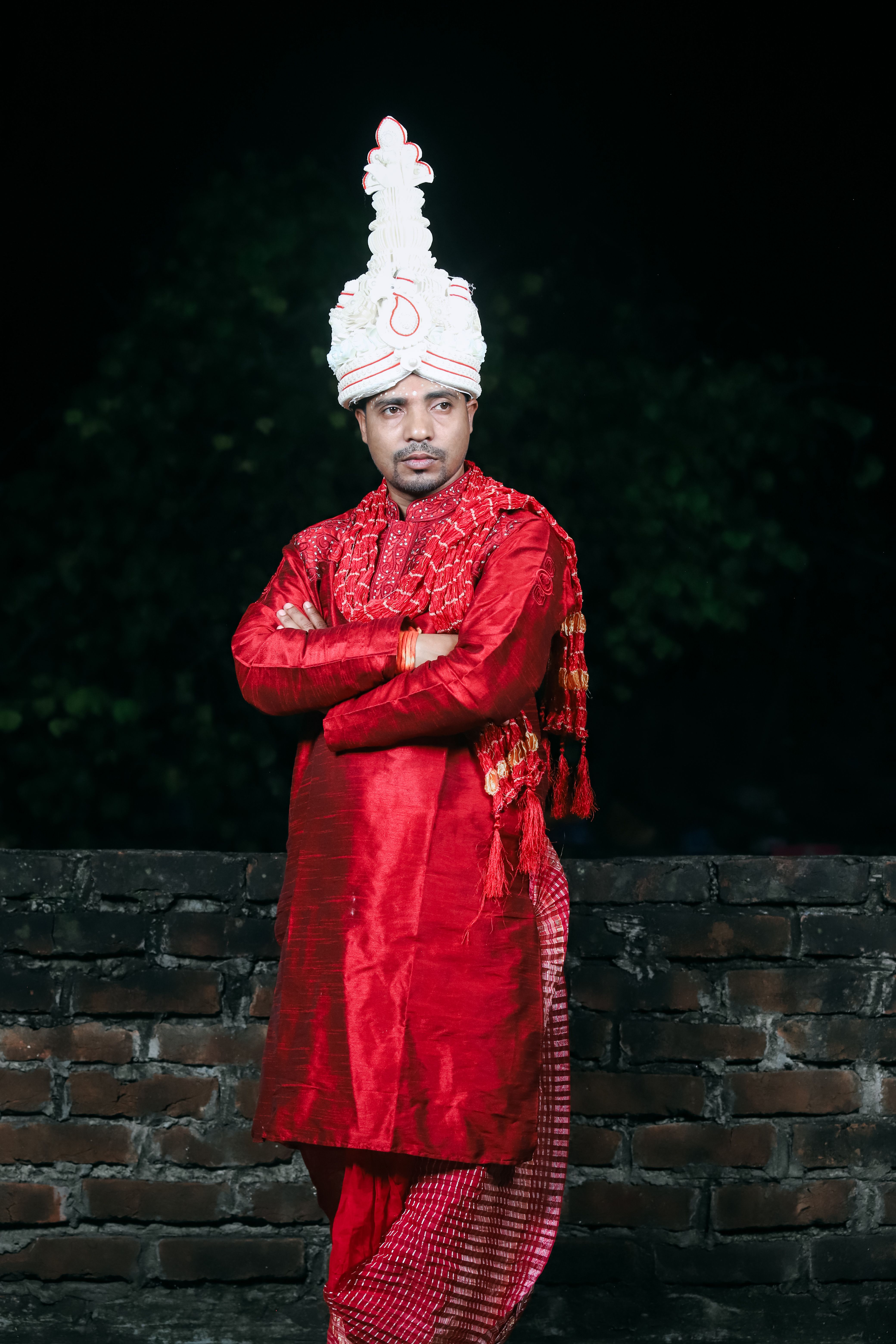 Portrait of a man in traditional red attire with ornate headgear, standing confidently.