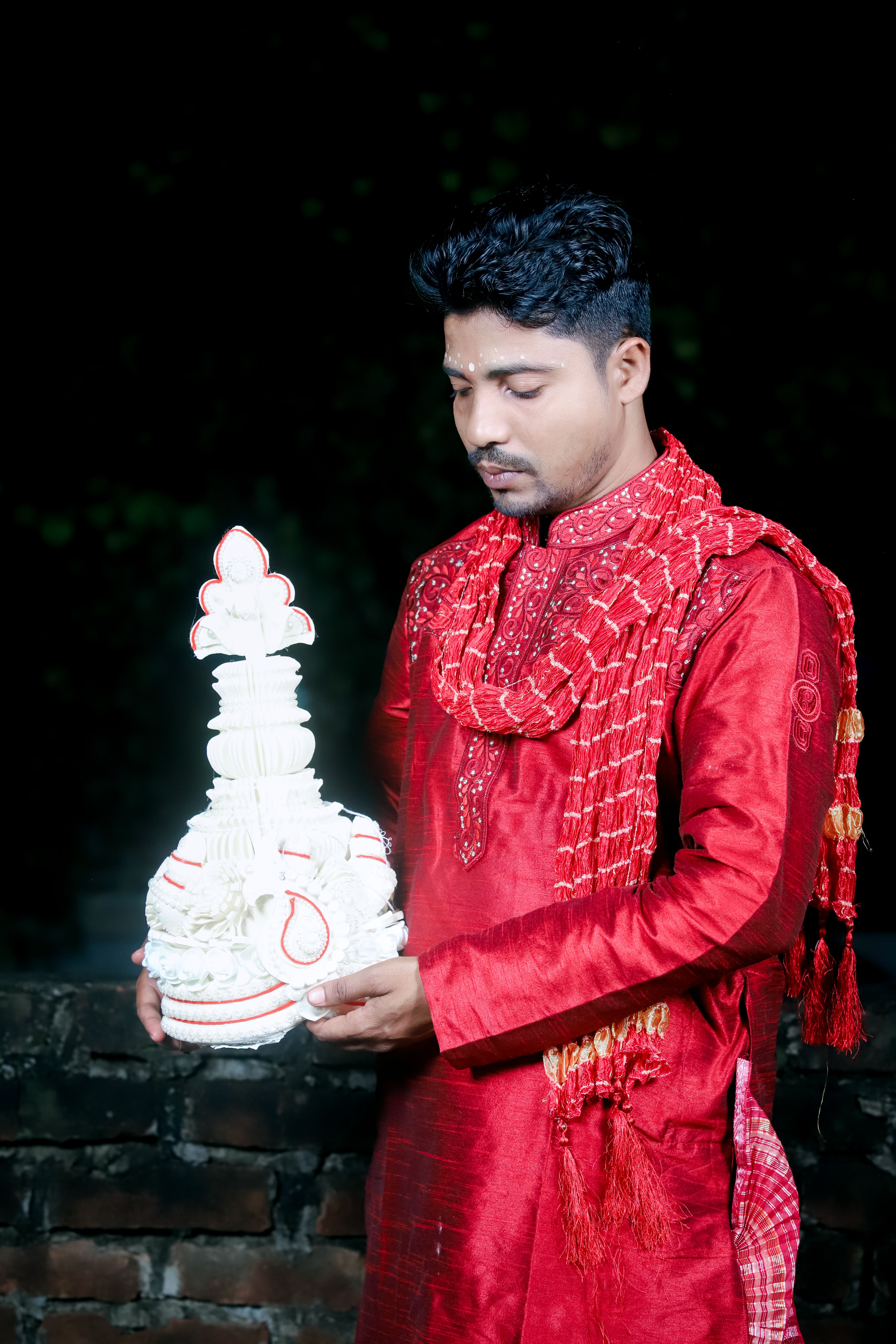 Man in vibrant red traditional attire holding ceremonial headwear outdoors at night.