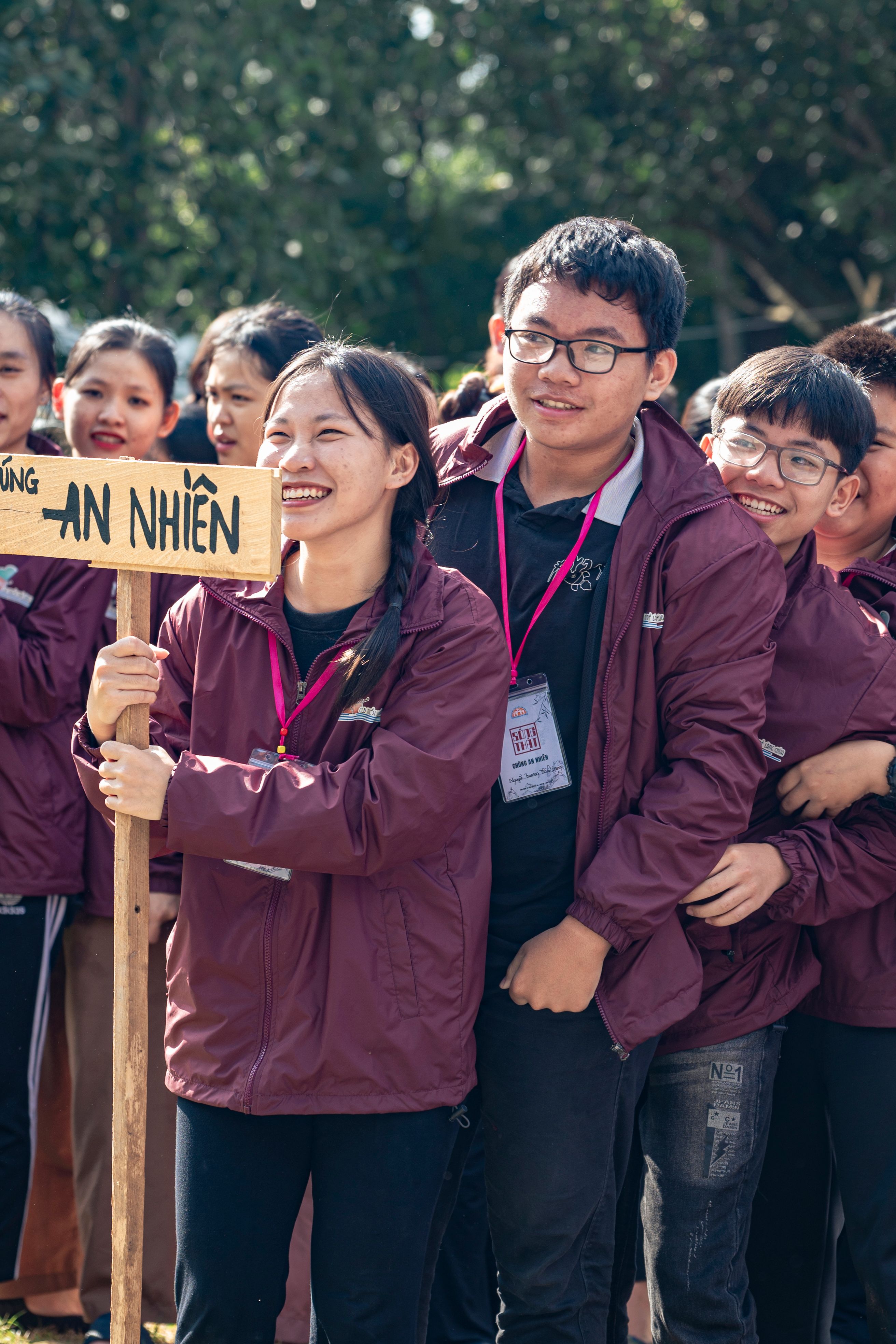 A lively group of young adults smiling and bonding at an outdoor event, holding a sign.