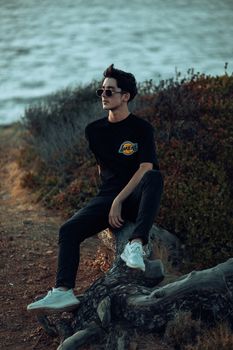 Fashionable young man sitting on a log by the lake during sunset, exuding a relaxed vibe.