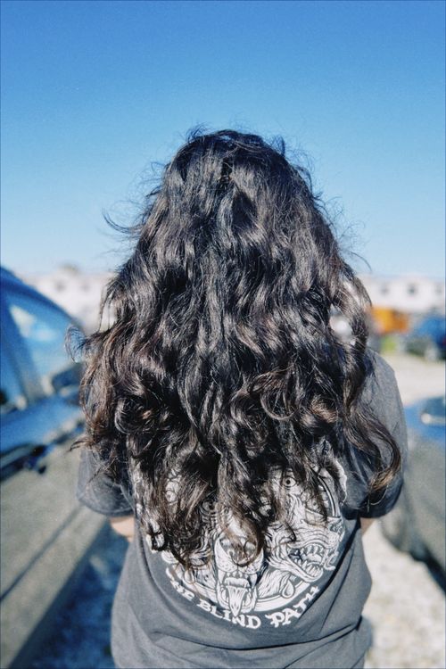 Free A woman with long curly hair stands outdoors, showcasing her hair against a clear blue sky. Stock Photo