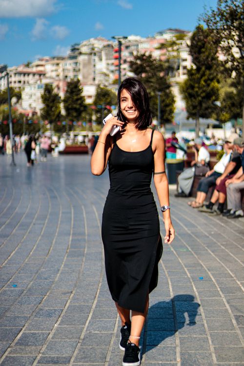 Free Young woman in a black dress walking confidently in a sunny urban plaza, exuding elegance and charm. Stock Photo