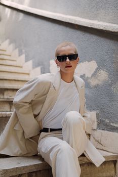 Chic woman in sunglasses and beige outfit sitting on stairs, modern urban fashion.