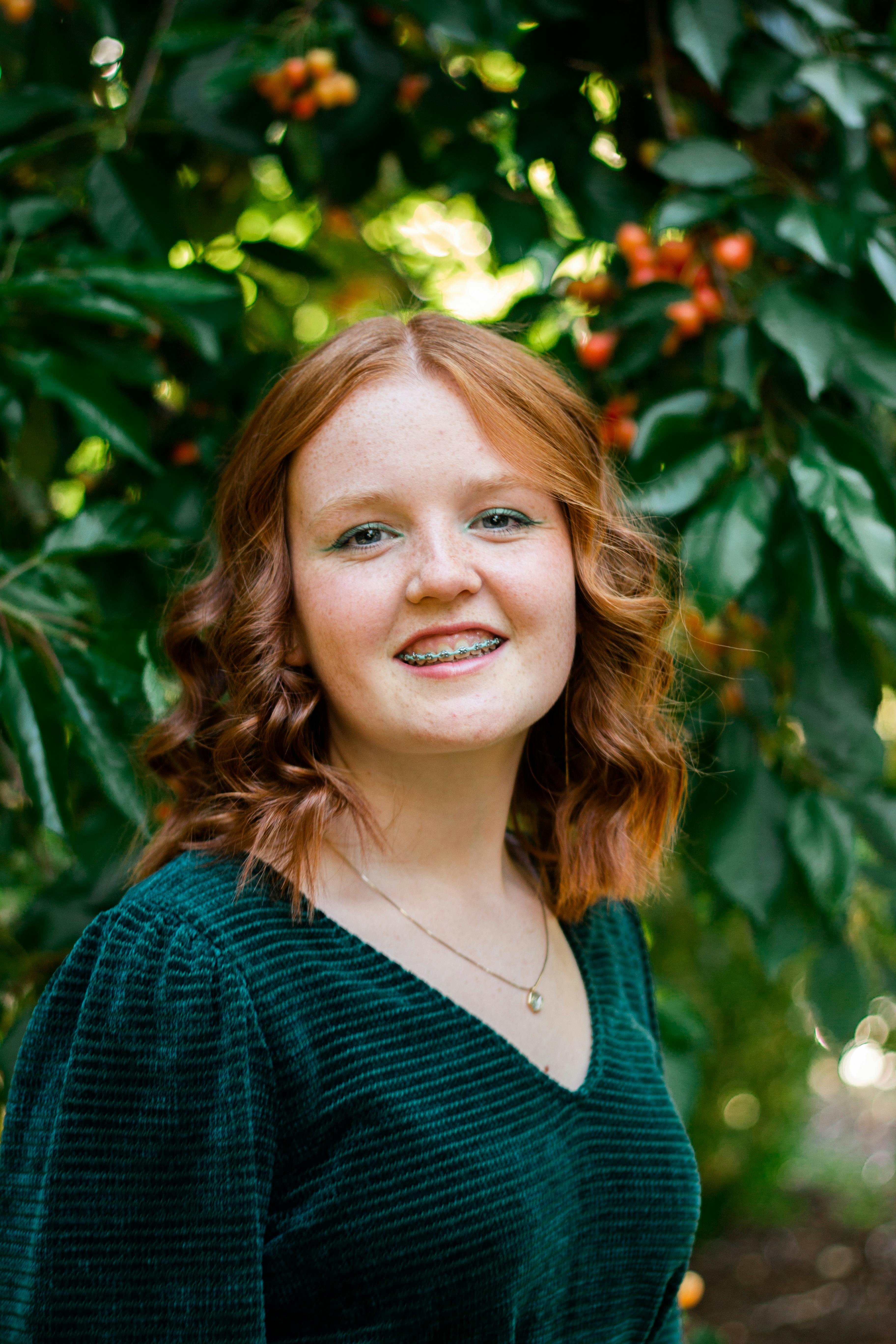 Red-haired woman smiling in an orchard with green dress and freckles, surrounded by foliage.