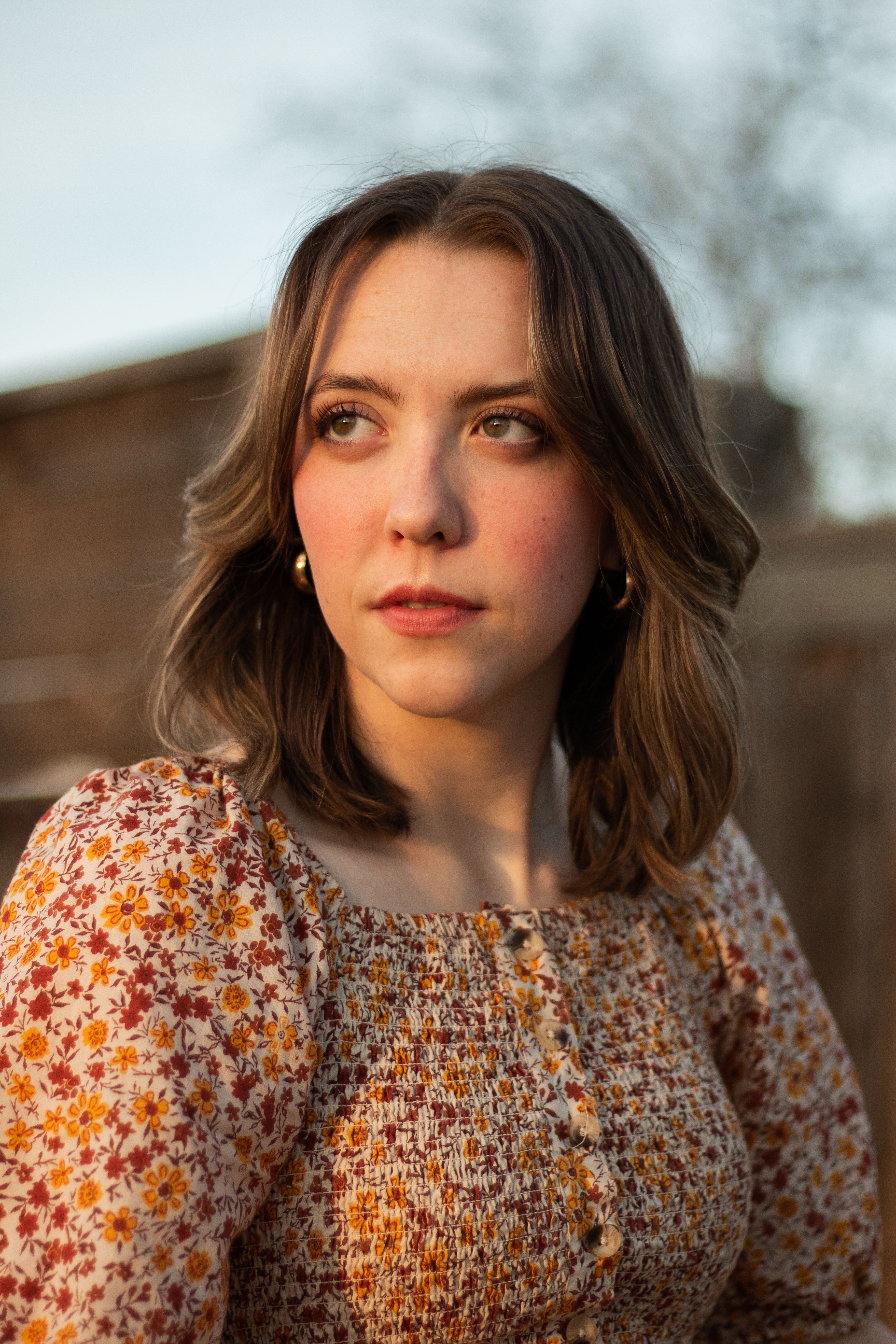 A young woman with brown hair poses outdoors in warm sunset lighting, wearing a floral dress.