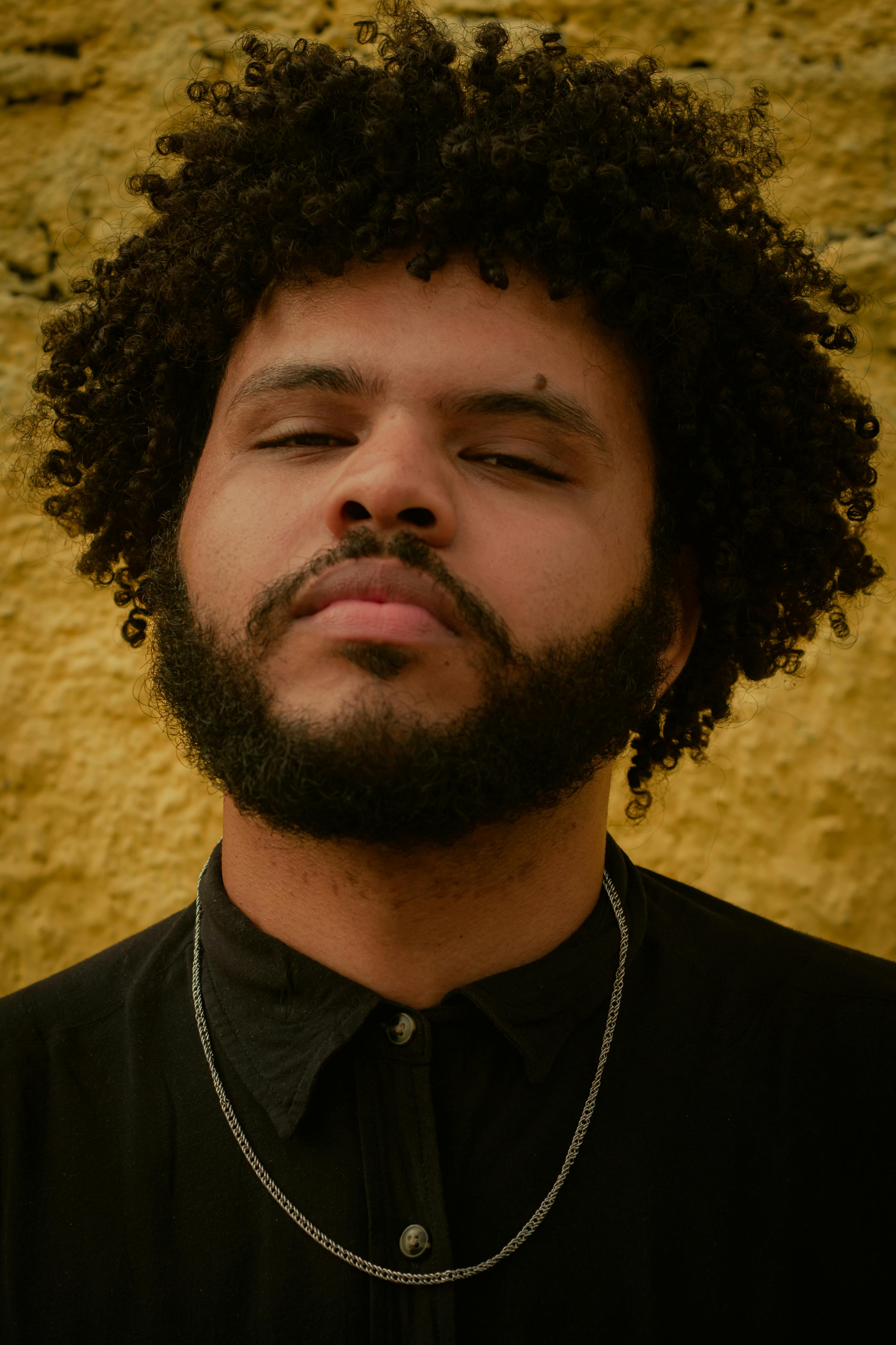 Close-up portrait of a man with curly hair and beard against a textured wall.