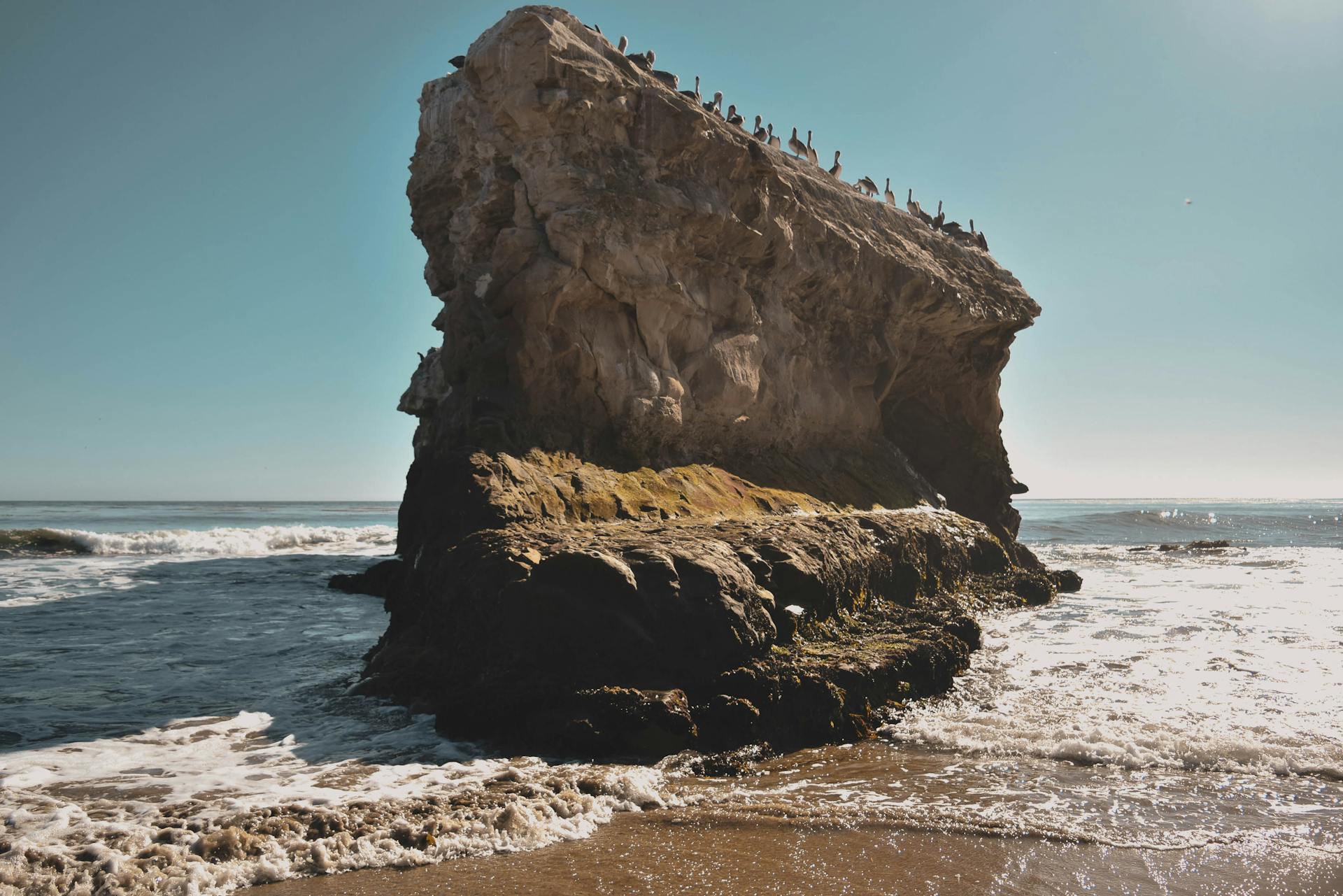Stunning rock formation at Santa Cruz with ocean waves crashing against it under a clear sky.