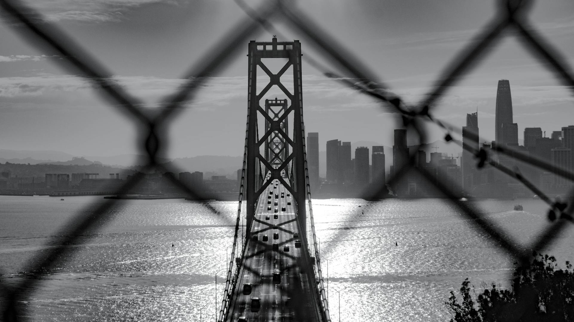 Black and white view of the Bay Bridge framed by a chainlink fence in San Francisco.