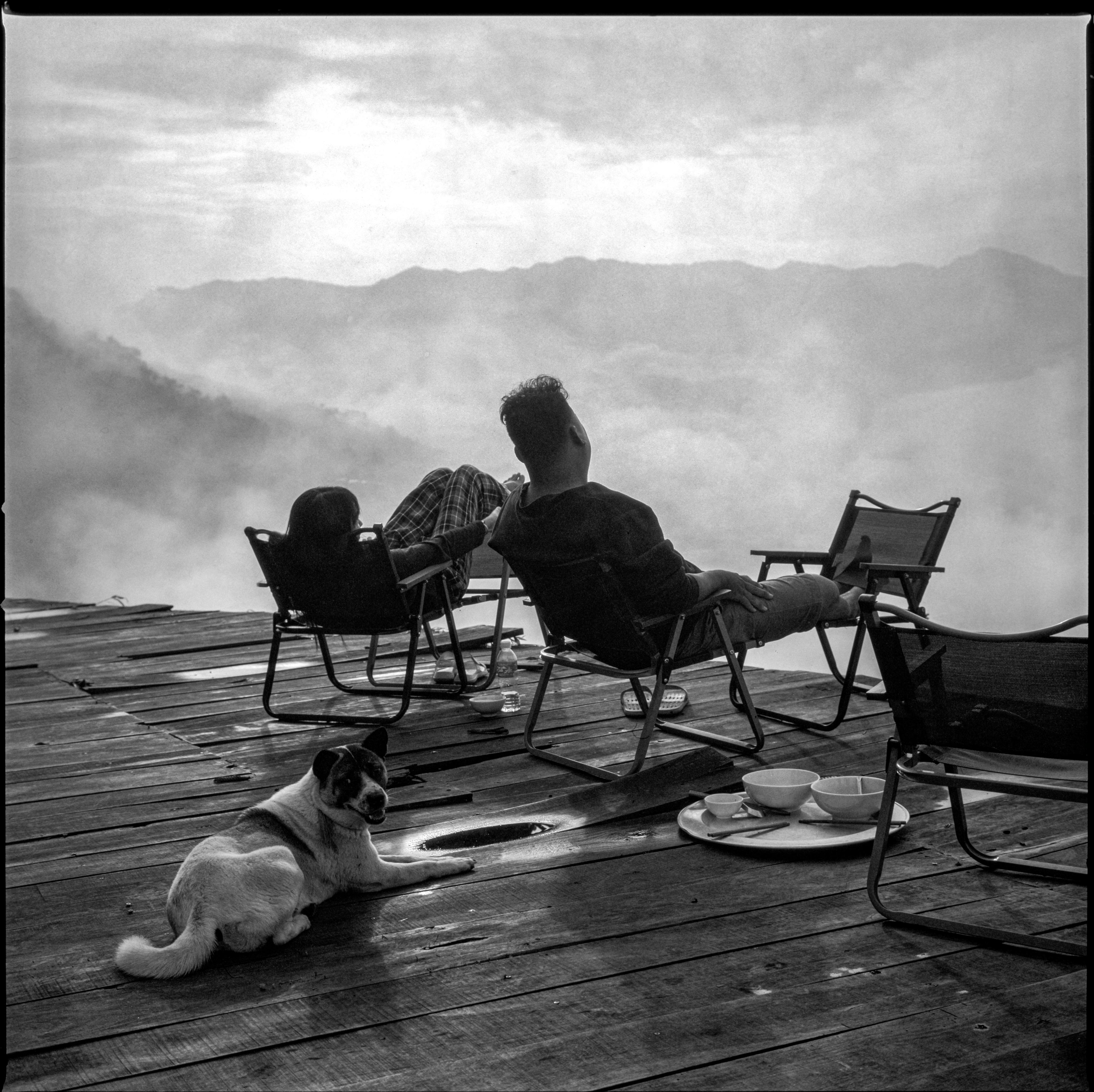 People relax on a wooden deck with chairs, a dog, and morning mist in Hà Giang, Vietnam.