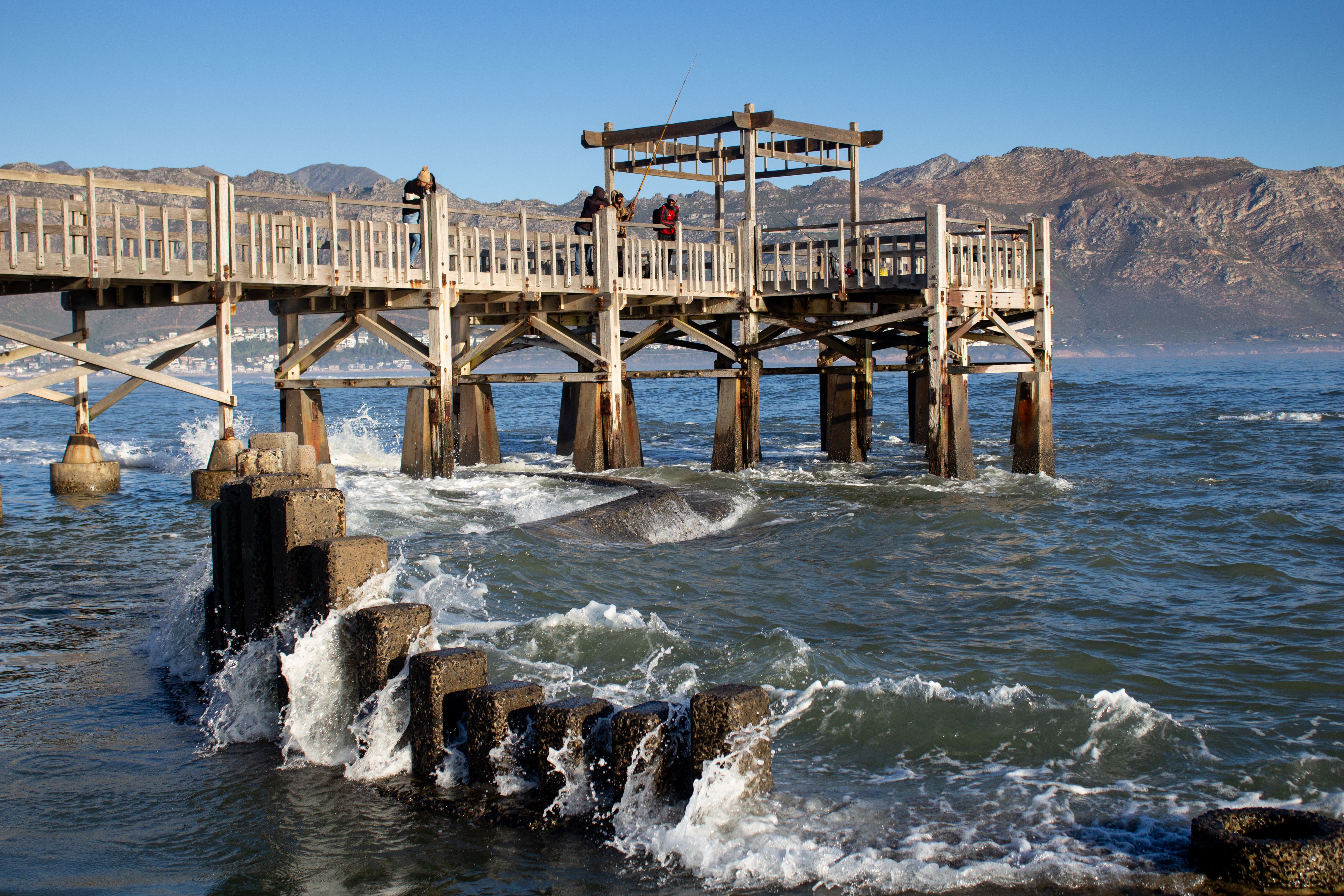A picturesque wooden pier extends over the ocean, framed by distant mountains under a clear sky.