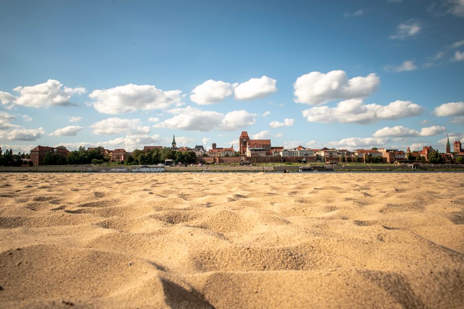 A scenic view of a sandy beach with a historic city skyline under a bright blue sky with clouds.