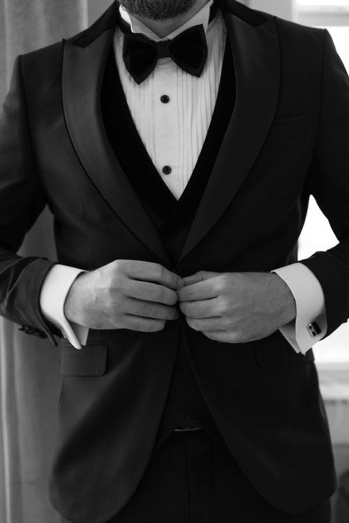 Free Black and white photo of a groom adjusting his bow tie in a formal tuxedo. Stock Photo