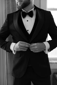 Black and white portrait of a man adjusting his formal tuxedo and bow tie, exuding classic elegance.