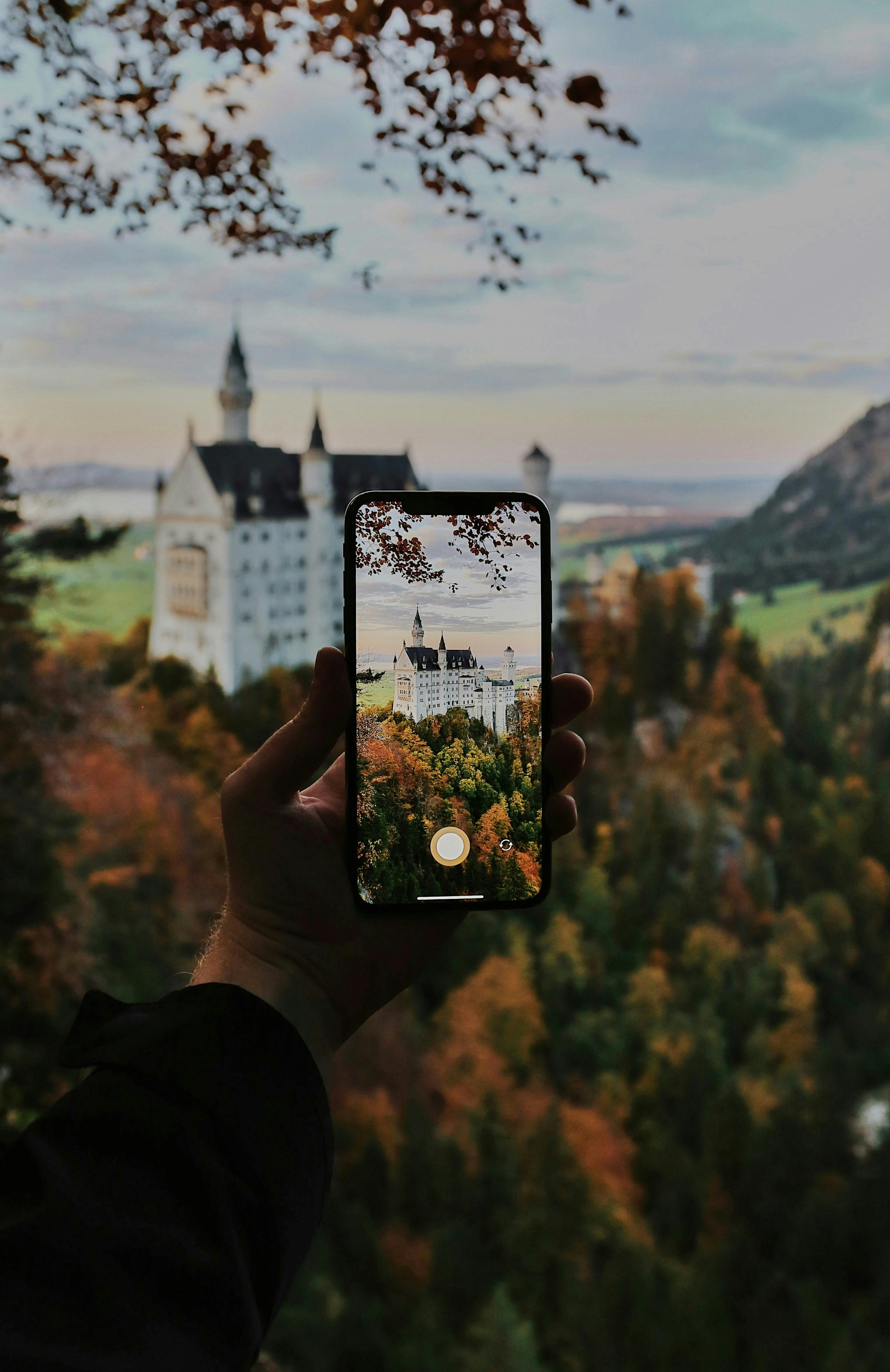 A smartphone captures the iconic Neuschwanstein Castle in autumn from a scenic viewpoint.