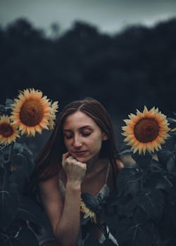A contemplative woman surrounded by vibrant sunflowers in a moody twilight setting.