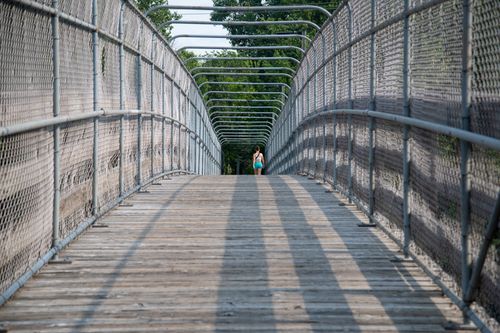 Free A woman walks on a wooden pedestrian bridge surrounded by metal railings in a summer setting. Stock Photo