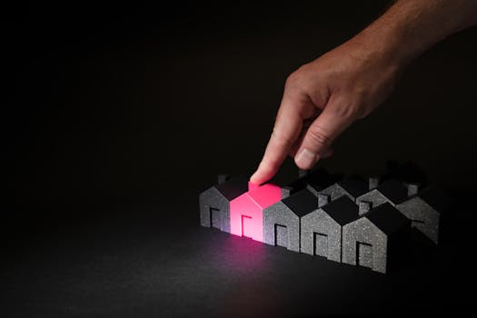 A hand selecting a glowing pink house among dark miniature houses, symbolizing real estate choices.