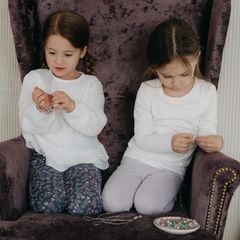 Two little girls in white jumpers on a plum velvet armchair threading beads, a dish of beads at their feet