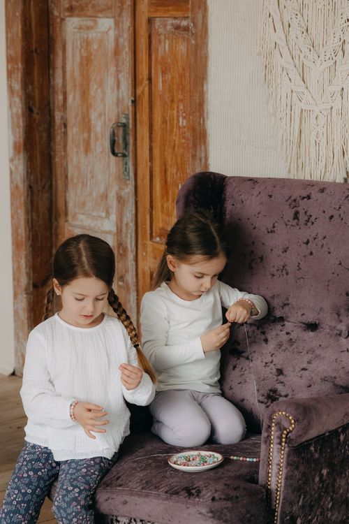 Free Two young girls sit indoors crafting beaded bracelets, showcasing creativity and family bonding. Stock Photo