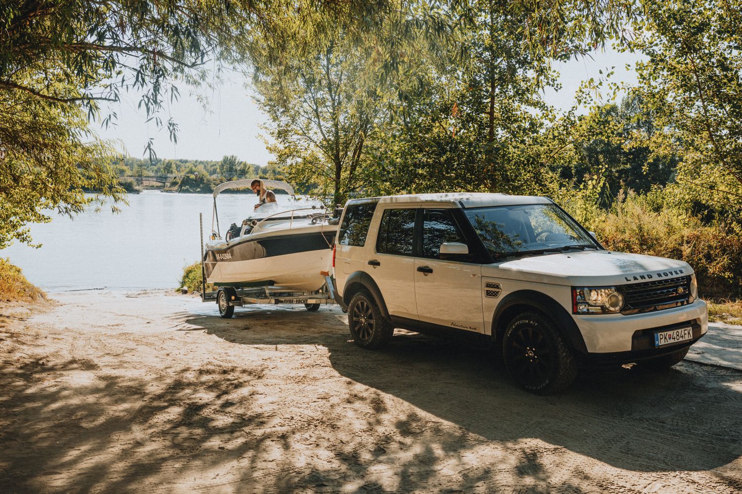 Truck towing a boat trailer down a sunlit road past a lake — trailer wiring and lights keep towing legal and safe