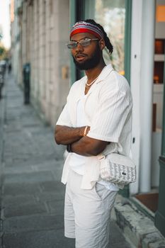 Fashionable adult male posing confidently in urban street setting, wearing sunglasses and carrying a bag.